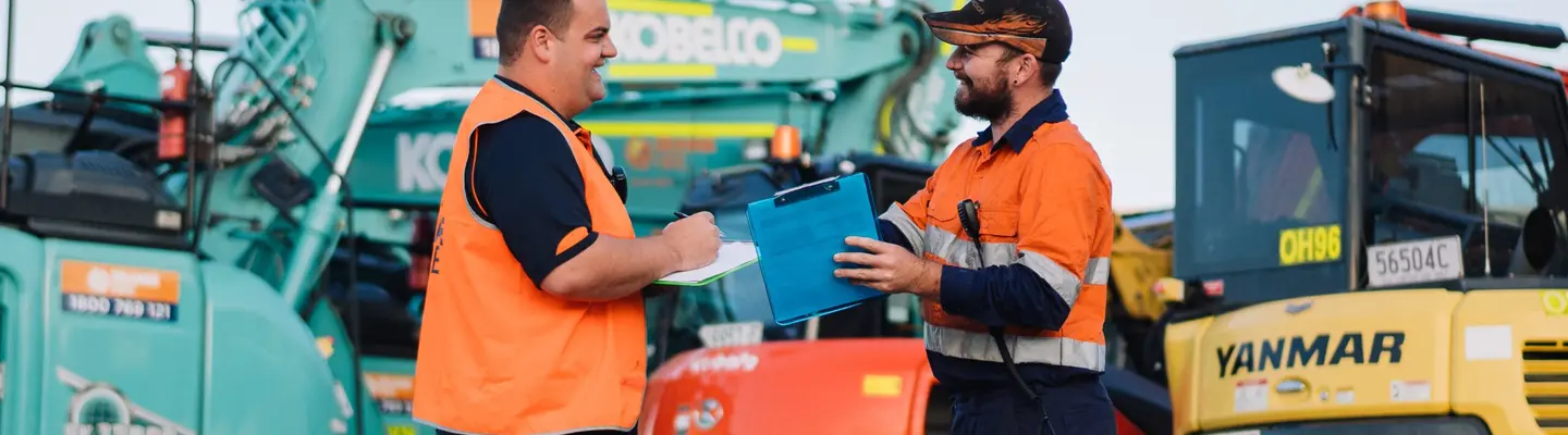 Deux travailleurs en uniforme souriant tout en se tenant devant l’équipement de construction sur un chantier.