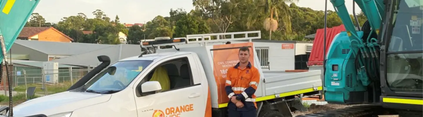 Homme debout à côté d’une excavatrice et d’un véhicule Orange Hire sur un chantier de construction avec des barrières de sécurité en place.