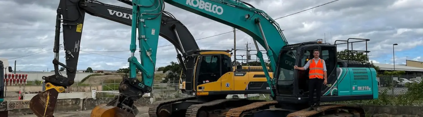 Homme debout dans une excavatrice stationnée sur un chantier de construction avec des barrières de sécurité en arrière-plan.