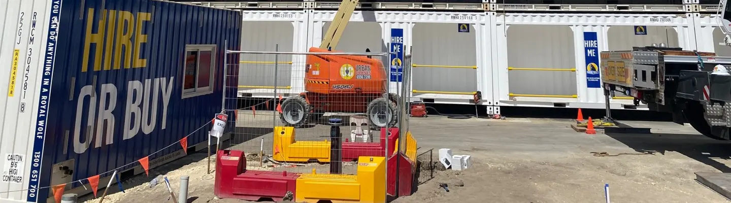 Site de construction avec conteneurs d’expédition utilisés comme bureaux et stockage, entouré de barrières de sécurité et de travailleurs en équipement haute visibilité.