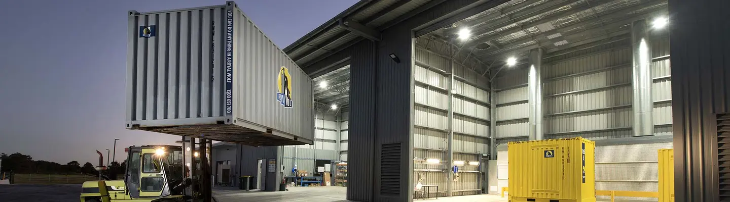 chariot élévateur soulevant un conteneur d’expédition à l’extérieur d’un grand entrepôt industriel avec un intérieur éclairé et des conteneurs supplémentaires à l’intérieur.