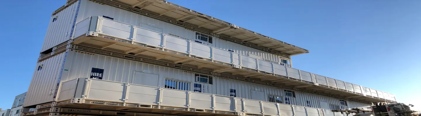 Une structure à plusieurs niveaux fabriquée à partir de conteneurs d’expédition blancs empilés, avec des balcons et des portes, installées à l’extérieur sous un ciel bleu clair, avec un affichage numérique du minuteur au premier plan.