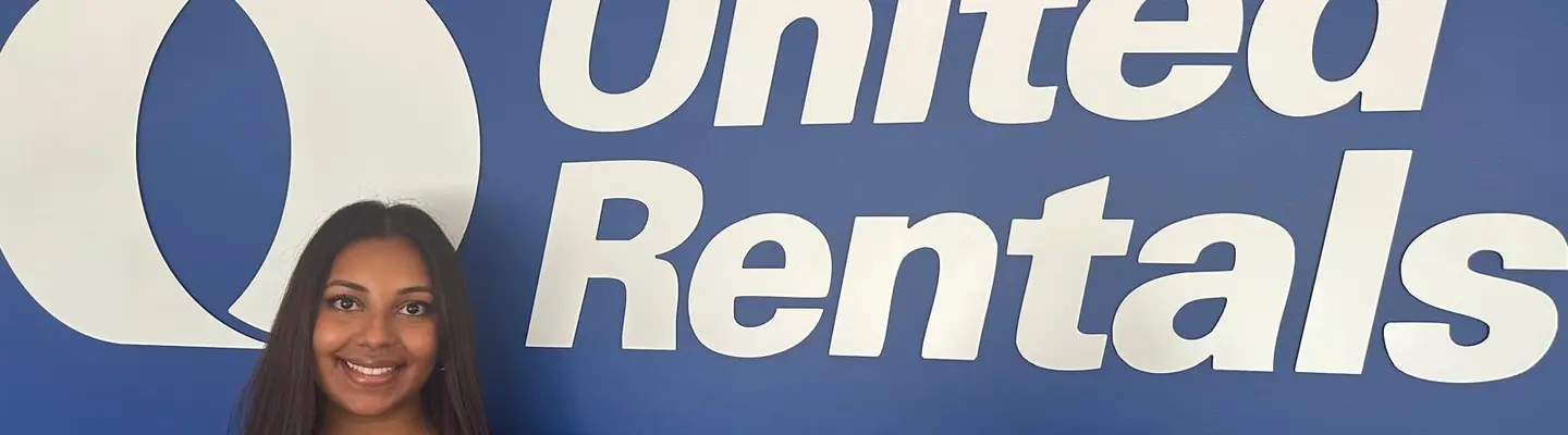 Une femme debout devant un mur bleu avec un grand logo United Rentals dessus.