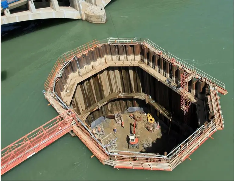 A large circular cofferdam made of interlocking sheet piles encloses an active excavation area with workers and equipment inside.