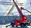 Vue latérale d’un chariot élévateur à portée variable Magni rouge avec flèche allongée, stabilisateurs, cabine fermée, travaillant sur un chantier de construction enneigé.