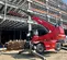 Vue avant d’un chariot élévateur à portée variable Magni rouge avec flèche et stabilisateurs allongés sur un chantier de construction devant un cadre en acier.