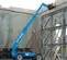 Vue latérale d’un chariot élévateur à portée variable Genie bleu avec flèche allongée et cabine fermée utilisée sur un chantier de construction.
