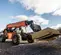 Un chariot élévateur à portée variable JLG SkyTrak orange et noir soulève une grande pile de contreplaqué sur ses fourches sur un chantier de construction, avec un bâtiment résidentiel visible en arrière-plan sous un ciel partiellement nuageux.