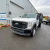 Un camion Ford blanc avec une plateforme à plateau devant un bâtiment industriel lumineux avec une porte de garage enroulable. Le camion est légèrement incliné vers la gauche, montrant l’avant et le côté droit.
