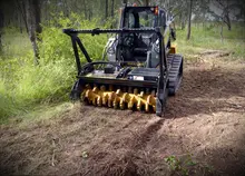 Large machine trimming overgrown grass in an open field near highway barriers to support roadside safety and visibility.