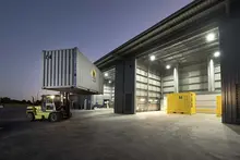 Forklift lifting a shipping container outside a large industrial warehouse with illuminated interior and additional containers inside.