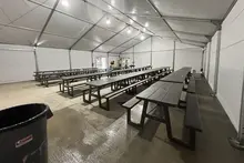 Rangées de tables de pique-nique sombres disposées à l’intérieur d’une tente industrielle spacieuse et lumineuse, servant de salle de pause temporaire.